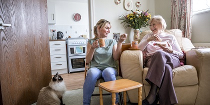 Senior woman sitting in her armchair listening to a younger woman talking. Both look happy as they enjoy a drink and snack together